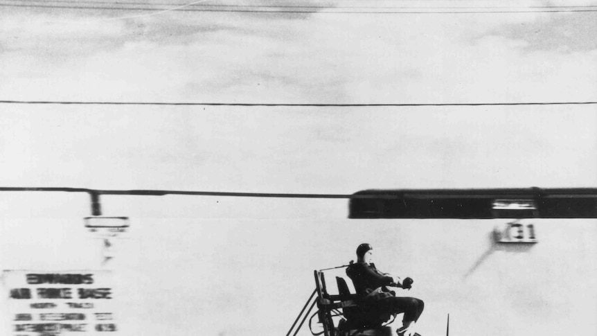 A man sits atop a sled-esque construction with rockets strapped to the back of it