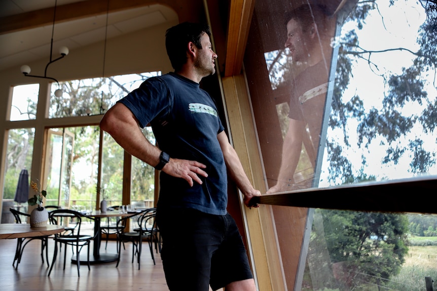 Man with black hair and white skin in blue t-shirt stands looking through large window with empty restaurant in background