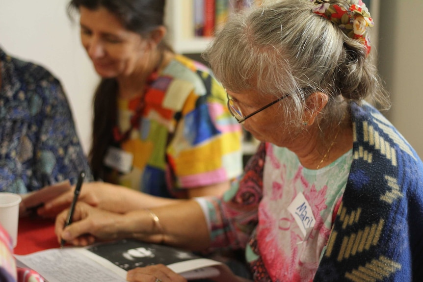 Focus on a middle-aged woman writing with another woman out of focus in the background.