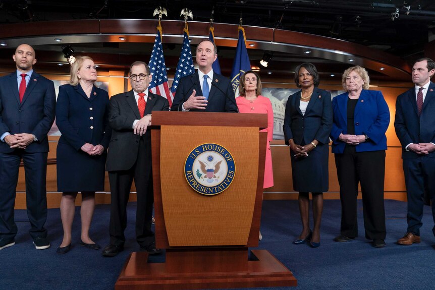 The seven impeachment managers and Nancy Pelosi stand on stage during a House briefing. Adam Schiff speaks from a podium.