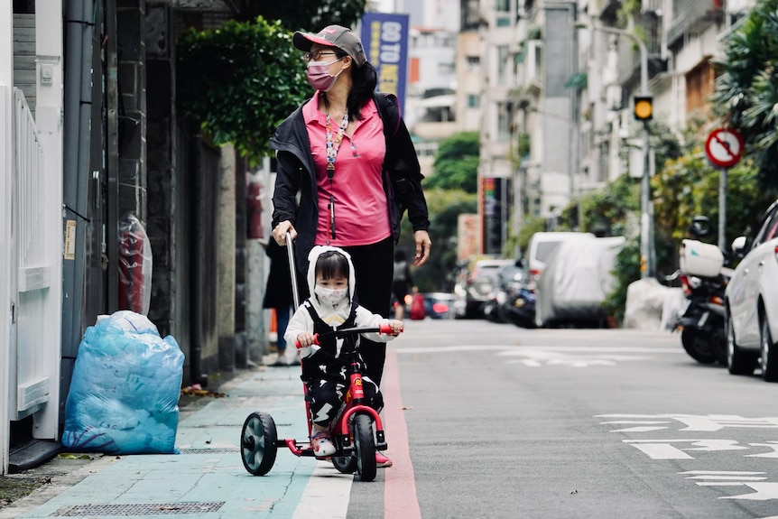 A woman wearing a pink mask and shirt walks down a street behind a toddler on a bike.