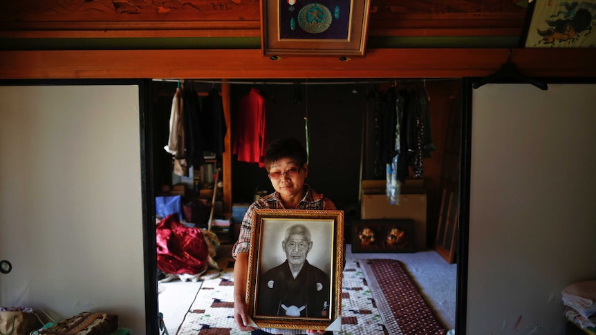 A middle-aged woman with Asian features holds a portrait of an older man with a similar ethnic make-up