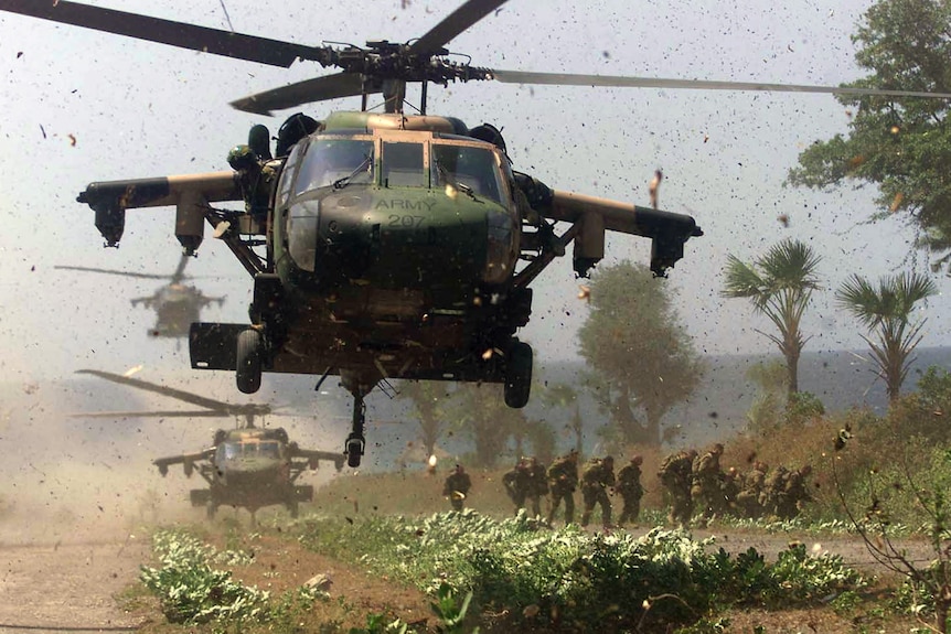 Three helicopters near the ground whip up dust and leaves. A group of soldiers walks away from one that has landed.