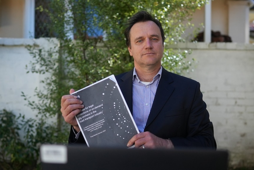 A middle-aged man in a collared shirt and suit jacket hold a report while looking serious.