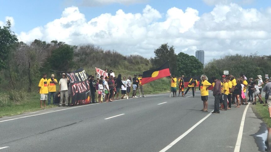 Indigenous protesters wearing yellow and carrying a large Aboriginal flag block a road.