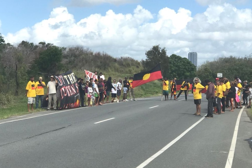 Indigenous protesters wearing yellow and carrying a large Aboriginal flag block a road.