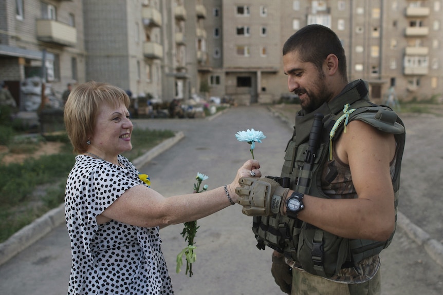 Женщина в платье в горошек улыбается, даря голубой цветок мужчине в военной форме.