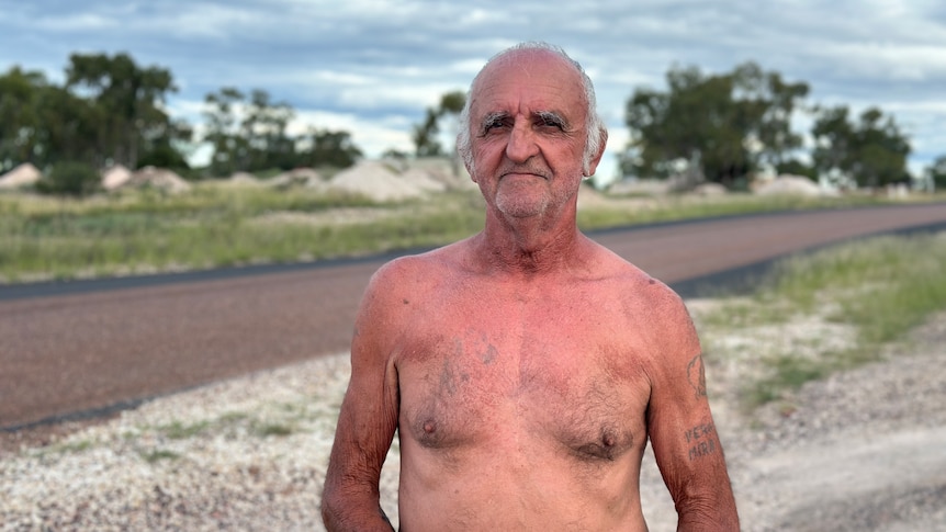 A older man without a shirt stands at the side of a road and looks at the camera with a serious face