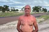 A older man without a shirt stands at the side of a road and looks at the camera with a serious face