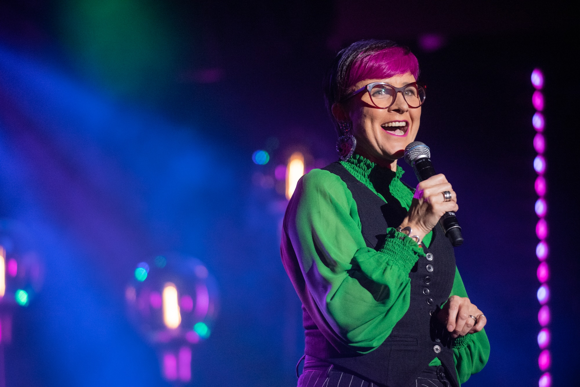 Cal Wilson wearing a green shirt, and with pink hair and glasses, holds up a microphone while smiling.
