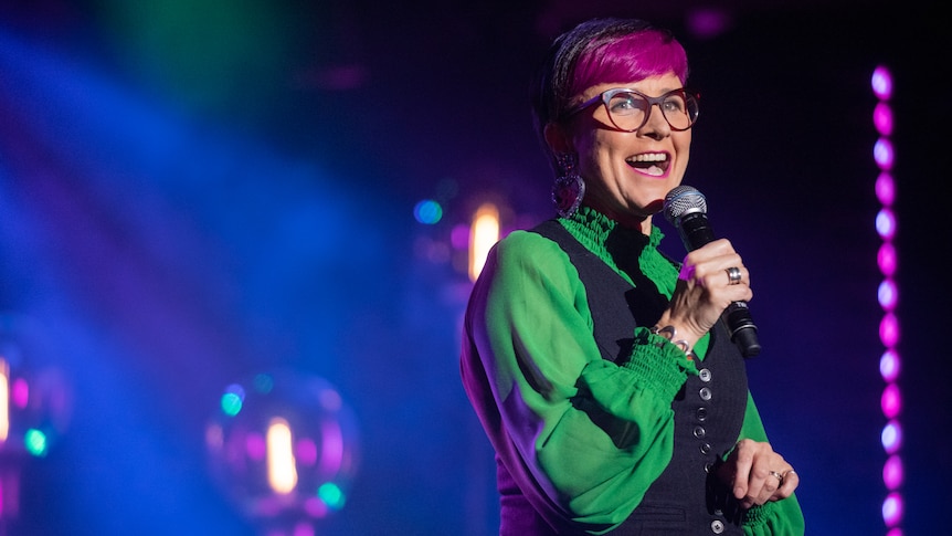 Cal Wilson wearing a green shirt, and with pink hair and glasses, holds up a microphone while smiling.