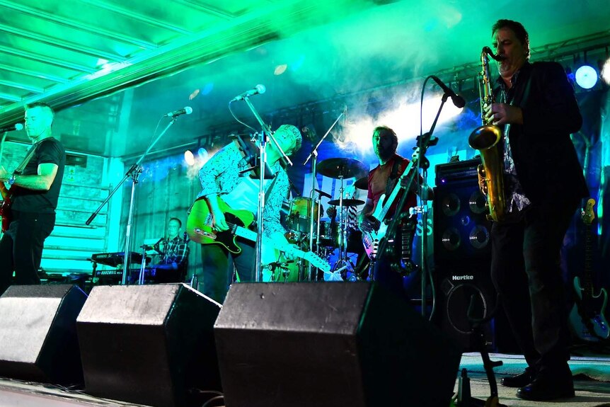 A band plays on a stage with green lighting and smoke effects.