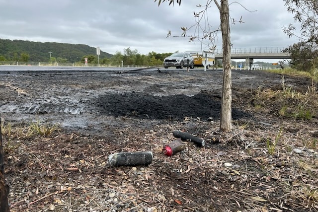 The burnt out remains of a fire on the side of the road where scorched aerosol cans lay on burnt grass
