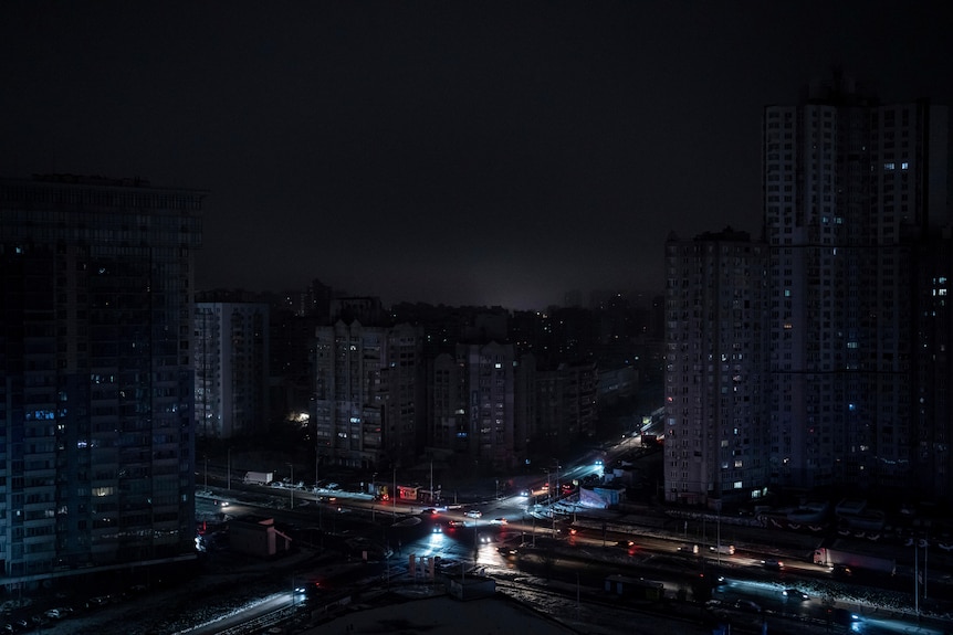 A wide photo at night of large buildings, mostly blacked out, with some cars on roads below with headlights shining.
