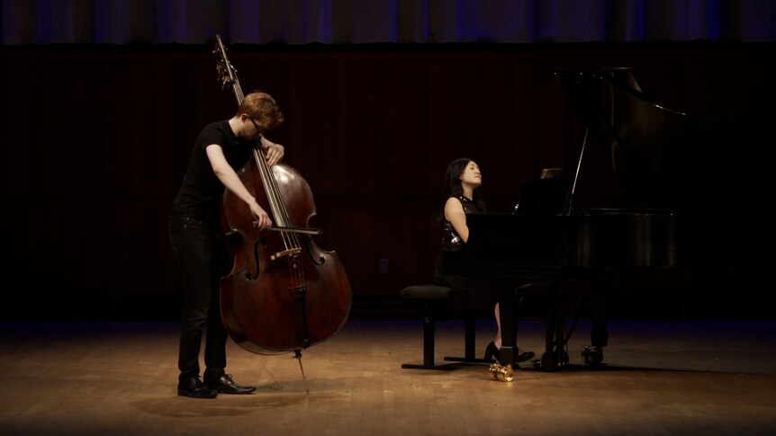 Jason Henery stands playing double bass on a dark stage with Jenny Chen on a grand piano in the background.