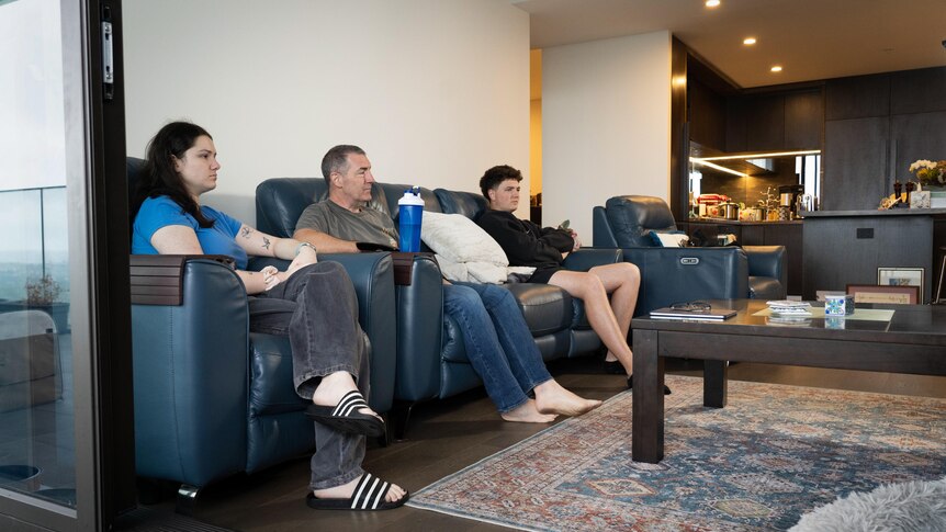 A father and his adult son sit on a lounge while his adult daughter sits on a couch inside their apartment.