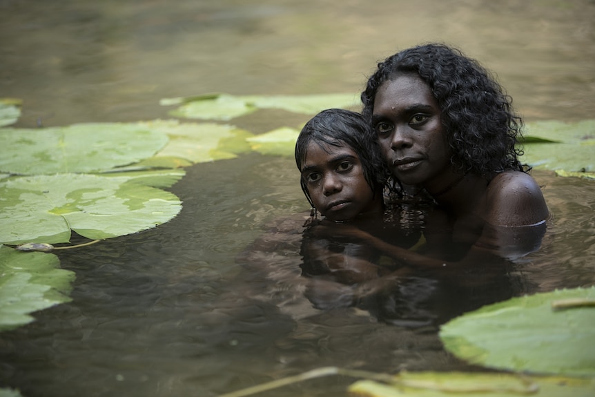 A still from the film High Ground of a young Aboriginal woman holding a boy in a river with lilly pads