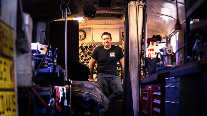 A man stands at the back of a bus which has been converted into a workshop and living space.