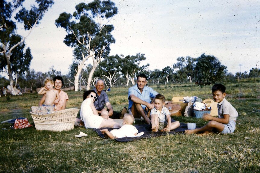 A family group sit on grass with trees in the background. The photo is from 1959 and shows adults and children.