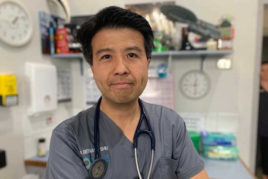 Dr Bernard Shiu wearing grey medical shirt with stethoscope over his shoulders standing in his consulting room