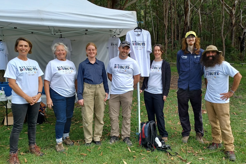 Group of 7 people standing on green grass with bushland behind