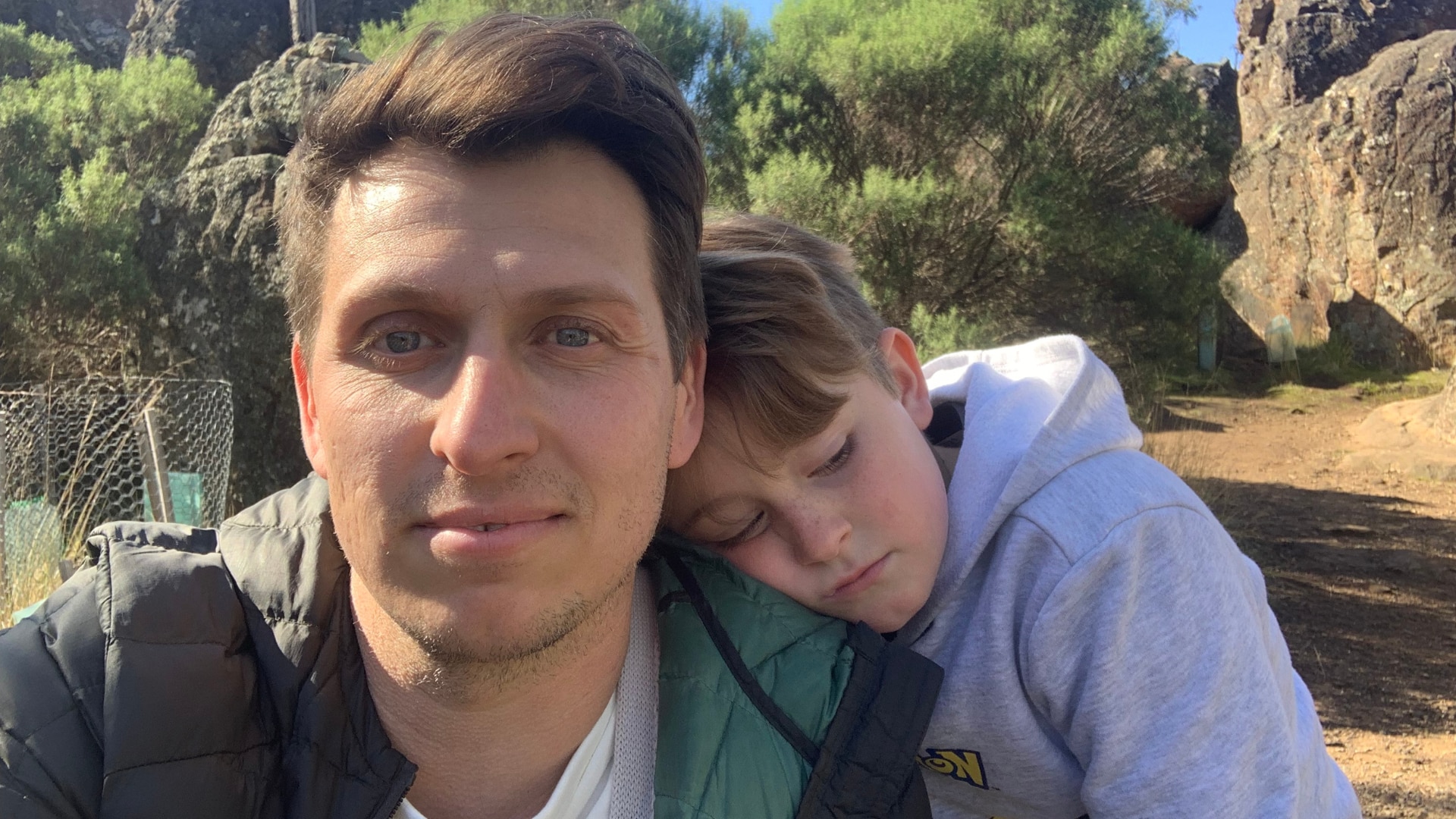 An eight year old boy leaning on the left shoulder of his father. Both look a little sombre.