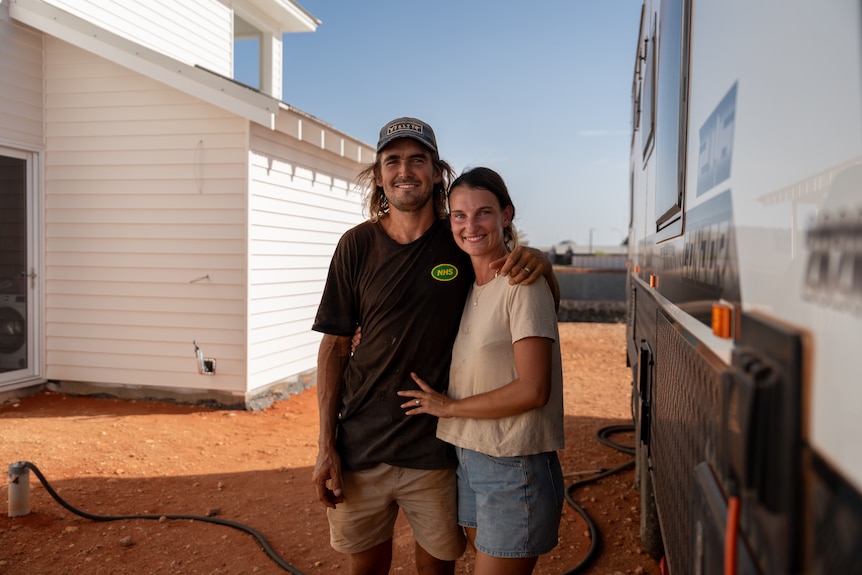 A man and a woman embracing next to a caravan and white house.