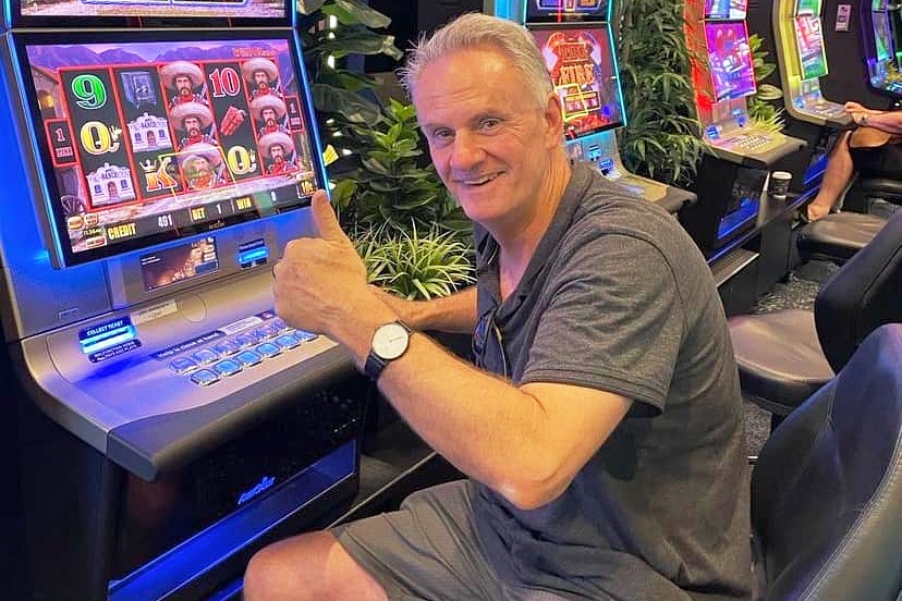 A man wearing a grey T-shirt and shorts, with his thumbs up, sitting in front of a poker machine inside a club.