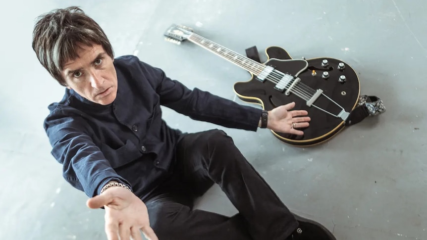 British musician Johnny Marr sits next to a black guitar with his arm outstretched