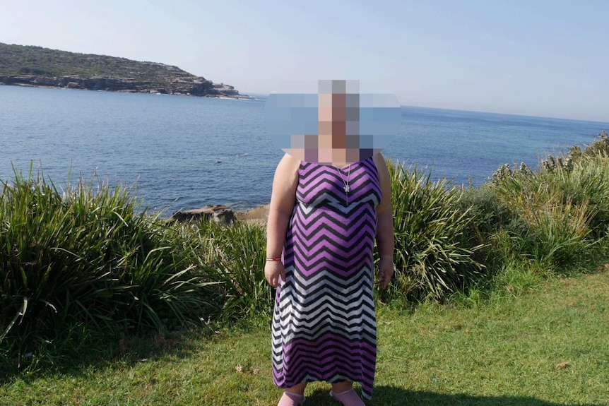 A woman wearing a purple dress stands in front of the ocean on a sunny day. She is unrecognisable as her face is pixelated.