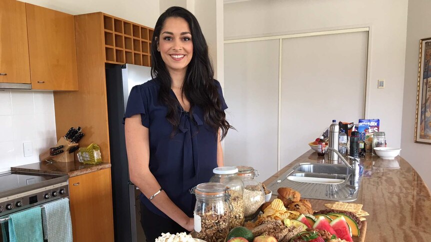 Dr Gina Cleo standing at a kitchen bench with food including fresh fruit, bread, cereal and popcorn.