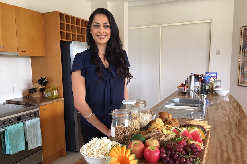 Dr Gina Cleo standing at a kitchen bench with food including fresh fruit, bread, cereal and popcorn.