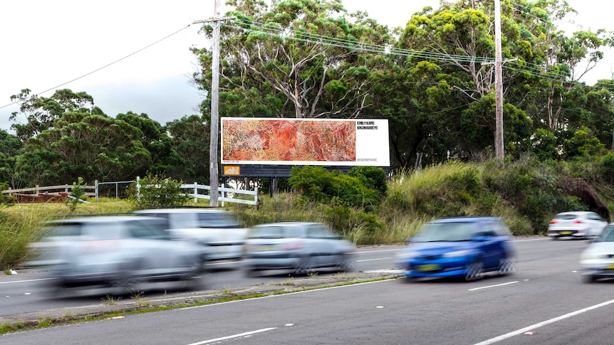 A billboard features a piece of art by a female artist on the side of a road as cars drive by.