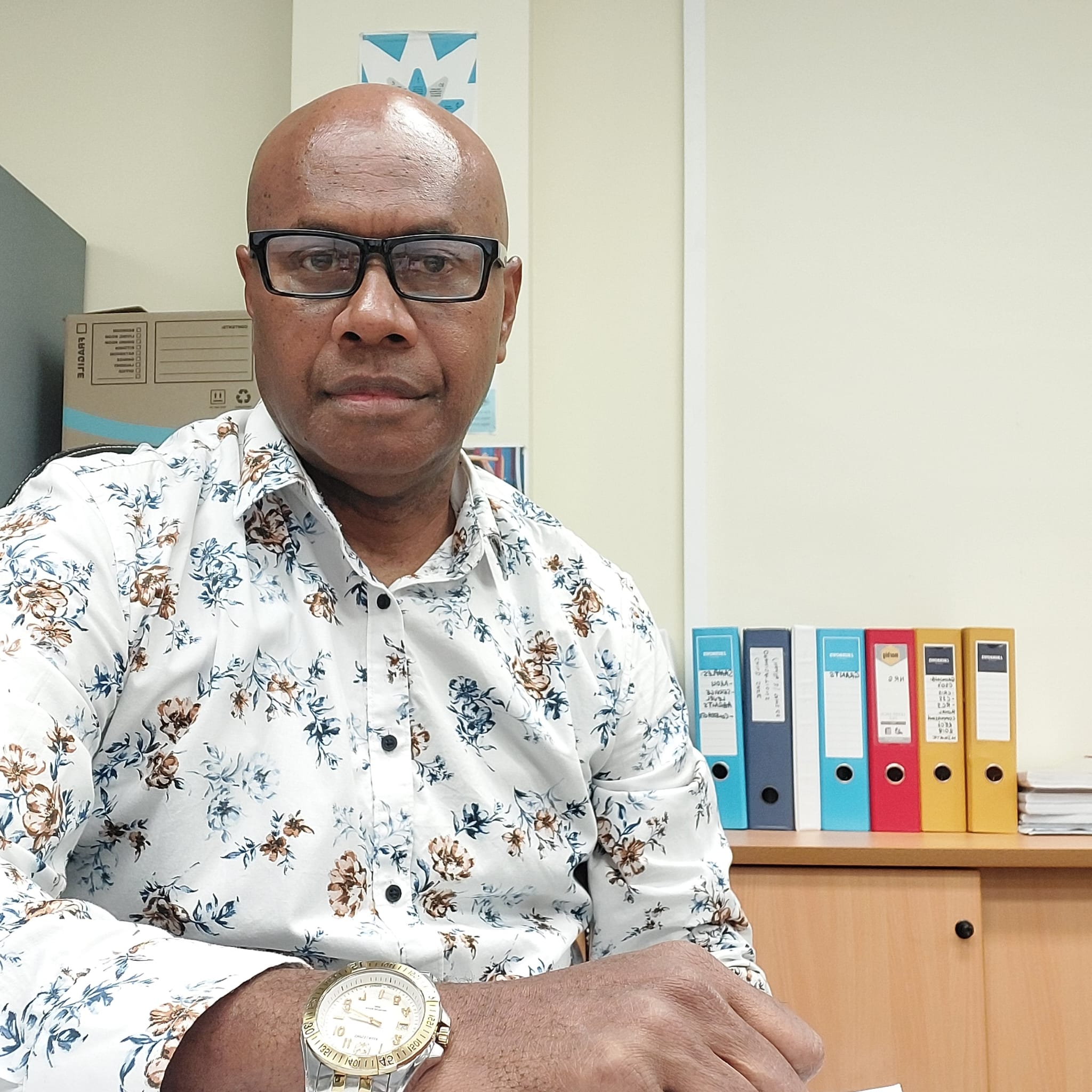 Neville Devete wears a floral collared shirt and looks seriously at the camera in an office.