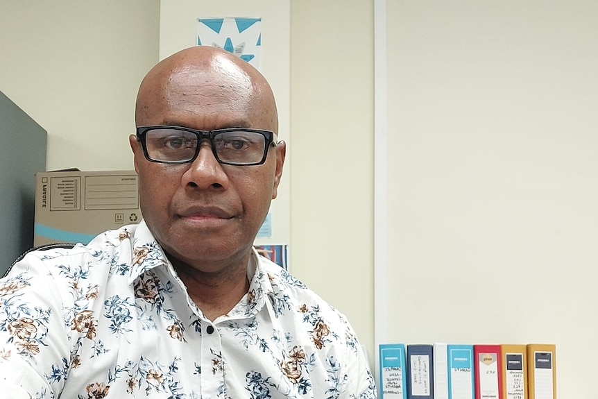 Neville Devete wears a floral collared shirt and looks seriously at the camera in an office.