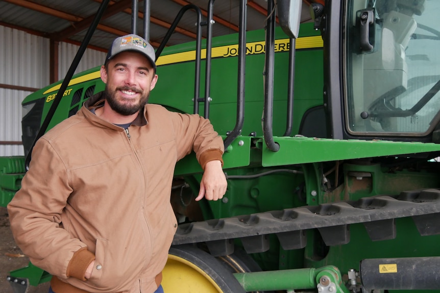 A man in a cap and brown jacket stands beside a green tractor.