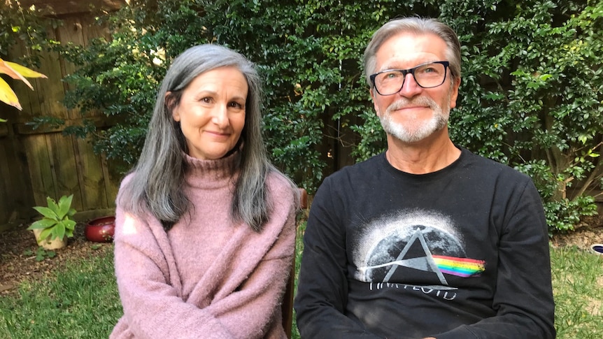 A woman in a pink jumper and a man in a Pink Floyd band shirt smile at the camera in a backyard.