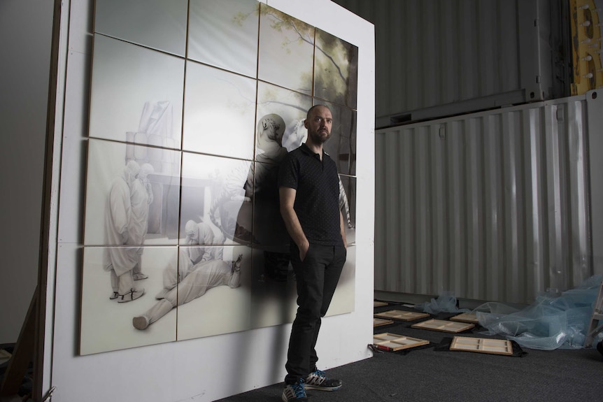 Sam Leach leans against his artwork in what appears to be a factory.