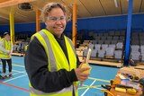 A girl wearing safety glasses and a fluoro yellow vest smiles at the camera while holding sandpaper and a piece of wood.