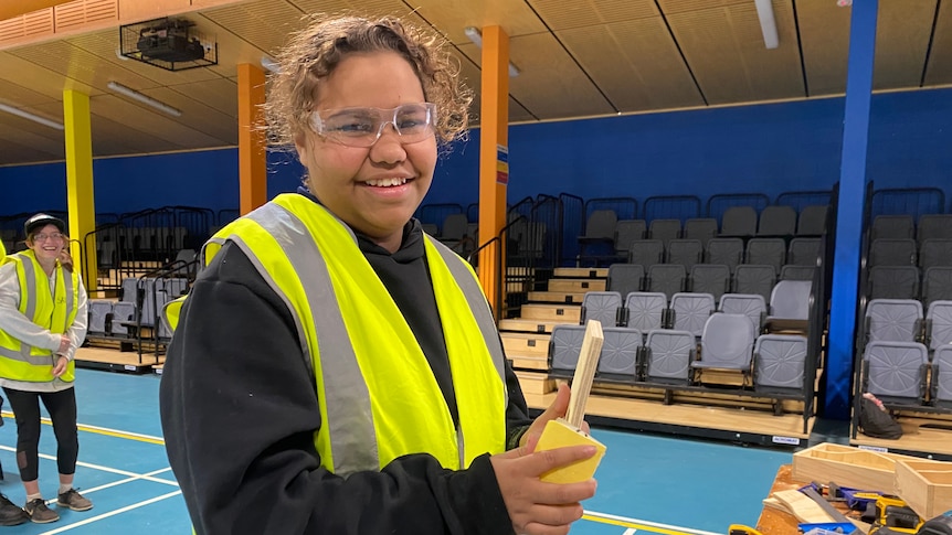 A girl wearing safety glasses and a fluoro yellow vest smiles at the camera while holding sandpaper and a piece of wood.