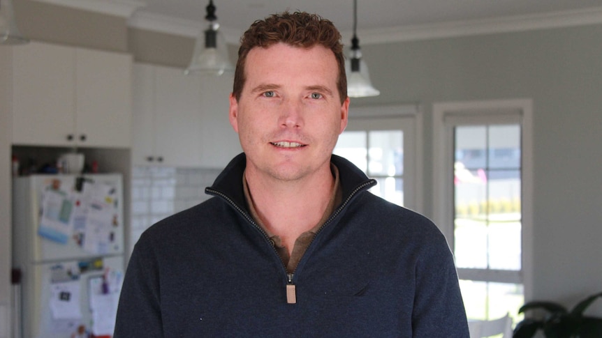 A young man with short hair stands in his kitchen, facing the camera.