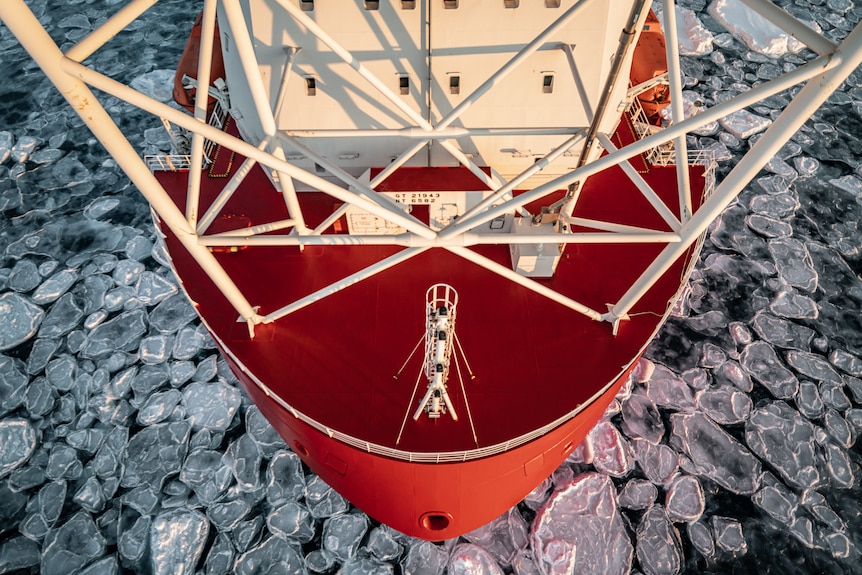 A large red ship with white beams at the top breaks through ice in the ocean.