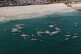 A drone shot of a large group of people gathering in boats and on surf boards in the ocean in a circle.