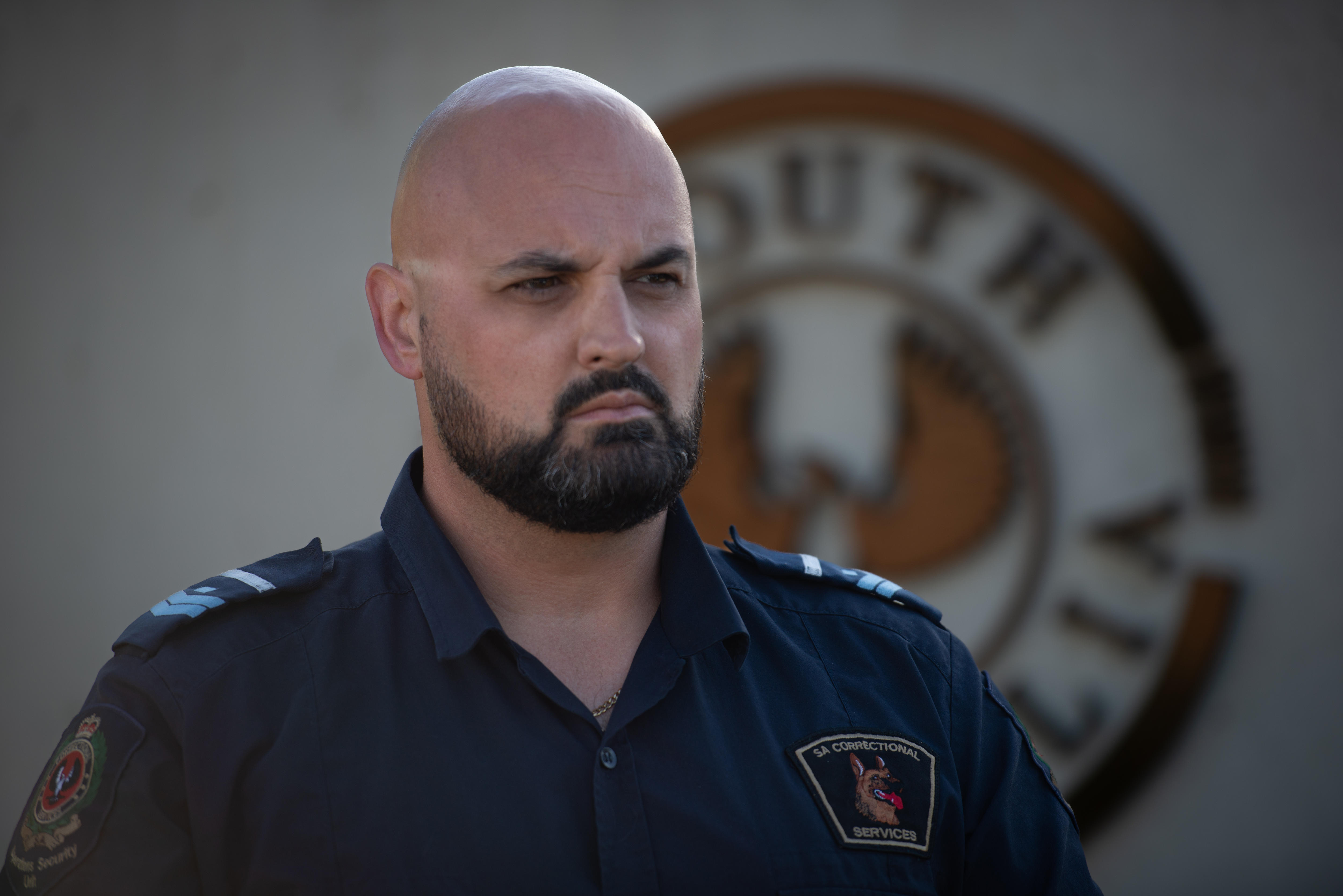 Un hombre con barba y uniforme naval de oficial de prisiones mira severamente frente a un muro de hormigón con el logotipo del gobierno de las SA.