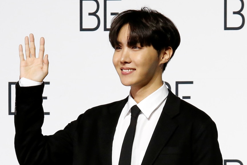 A young korean man in a suit waves to someone off camera.  