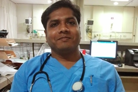 A man dressed in blue scrubs smiles as he sits on a chair in a hospital room.