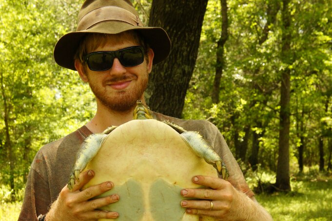 Man stands in the bush holding a turtle smiling at the camera.