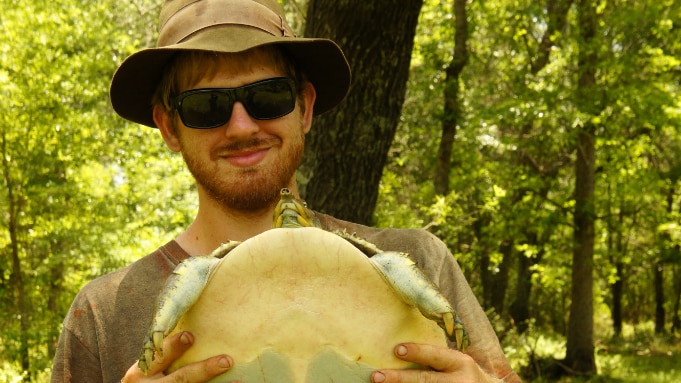 Man stands in the bush holding a turtle smiling at the camera.