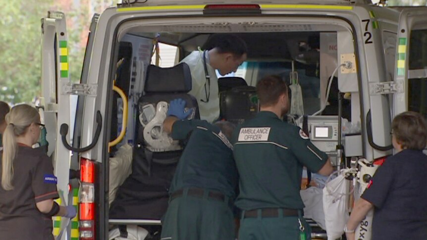 An ambulance sits parked with its back doors open and four medical staff standing behind it.
