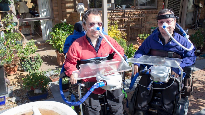 Two men with ventilators sit in wheelchairs outside a brick house, surrounded by plants.